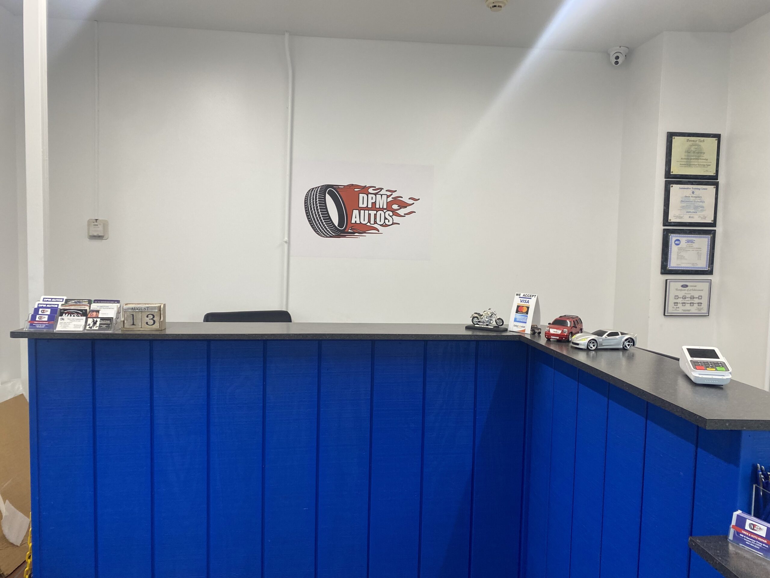 Bright blue reception desk at DPM Autos, featuring toy cars, brochures, a payment terminal, and framed certificates on the wall.